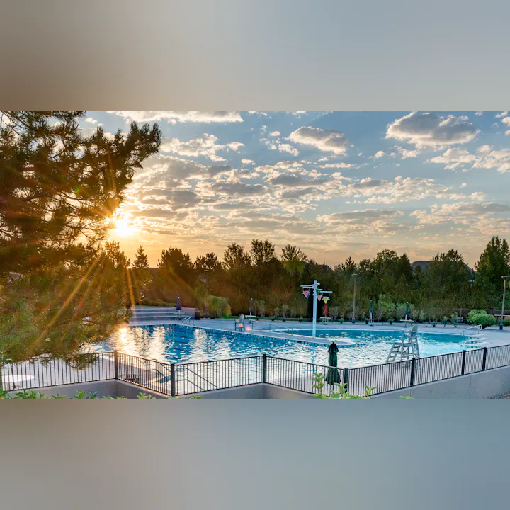A scenic view of a swimming pool at sunset, surrounded by trees and benches, with a clear sky and clouds.