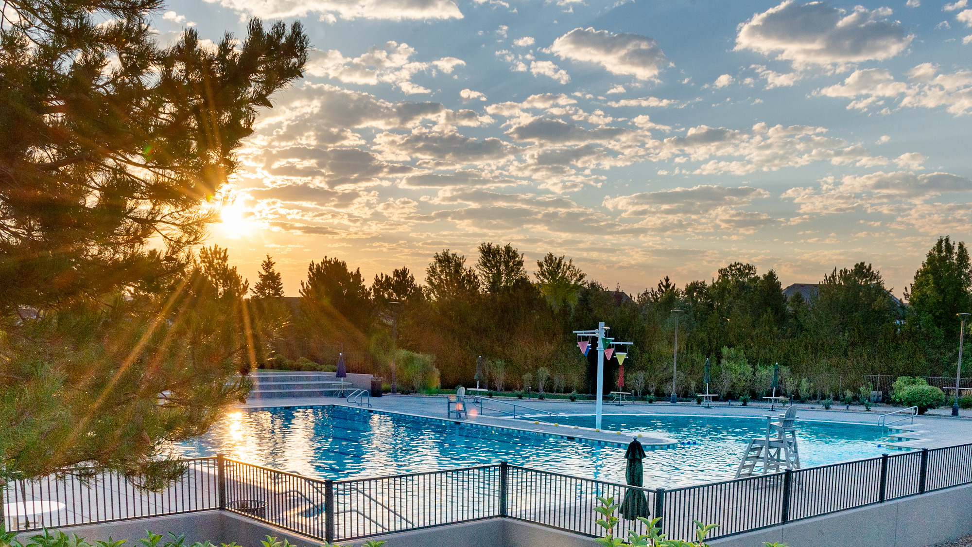 A scenic view of a swimming pool at sunset, surrounded by trees and benches, with a clear sky and clouds.