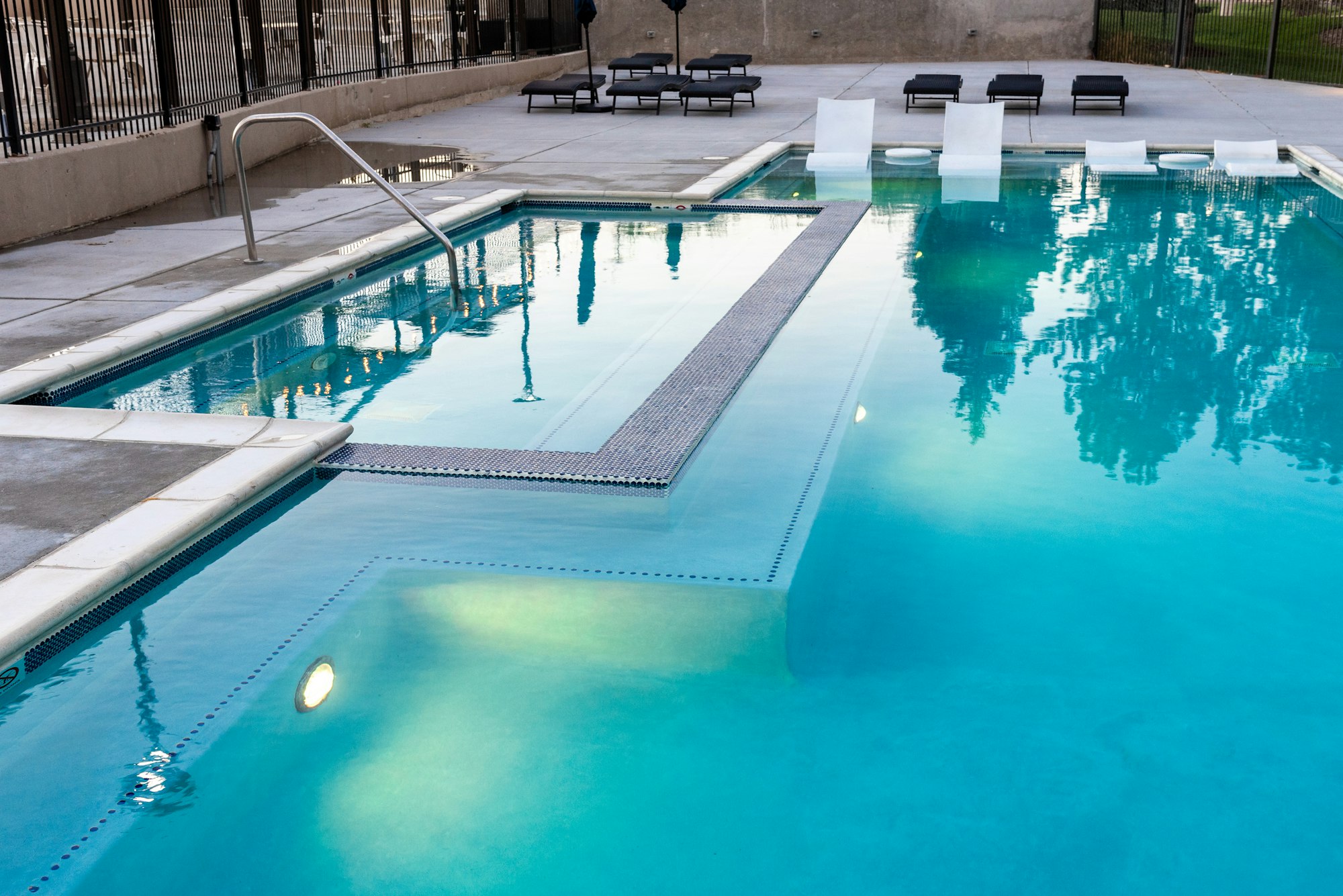 A clear swimming pool with a step, metal railing, and lounge chairs nearby, reflecting surrounding greenery.