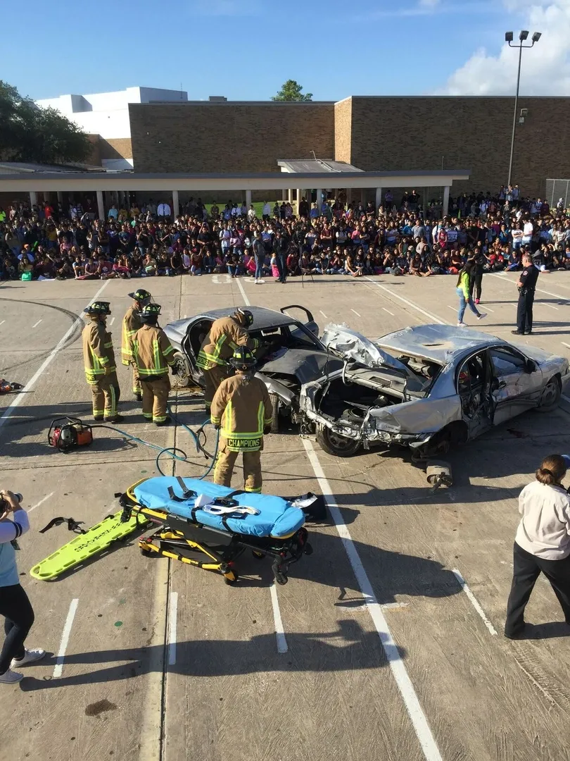 Firefighters demonstrate a car crash response in front of a crowd, with damaged cars and a stretcher on scene.