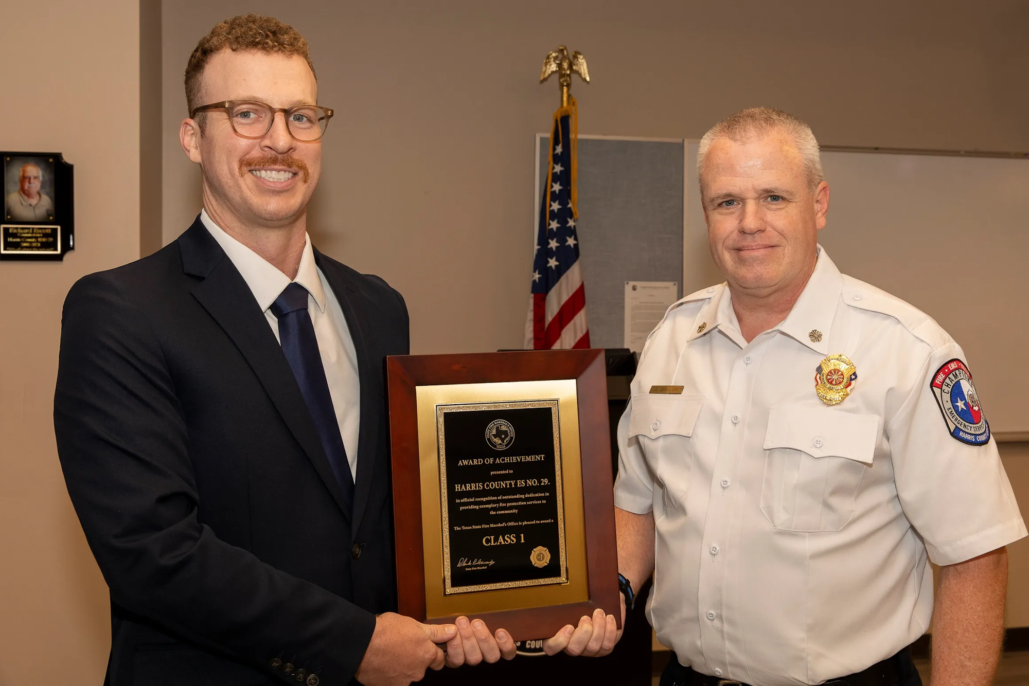 Two men holding an award. One is in a suit, the other in a uniform. An American flag is in the background.