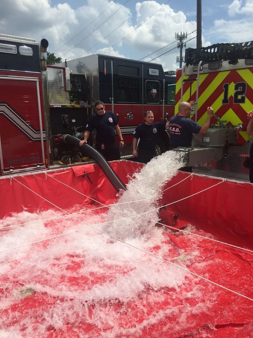 Firefighters fill a large red portable tank with water using a hose from a fire truck.