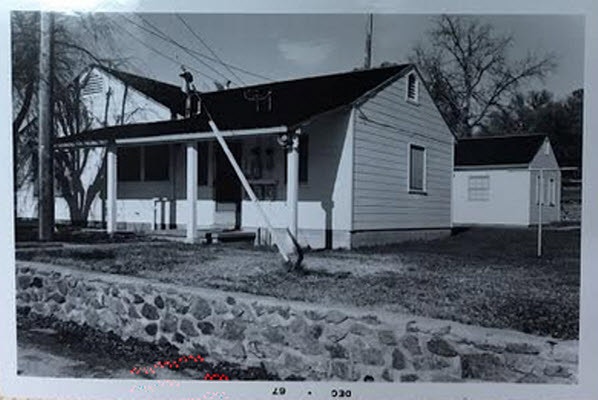 Black and white photo of a traditional house with a small front porch and a stone wall in front.