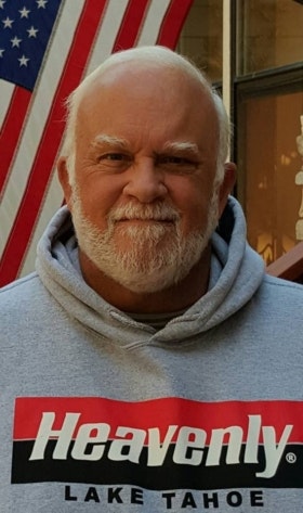 A smiling man with a beard wearing a "Heavenly Lake Tahoe" hoodie with an American flag in the background.