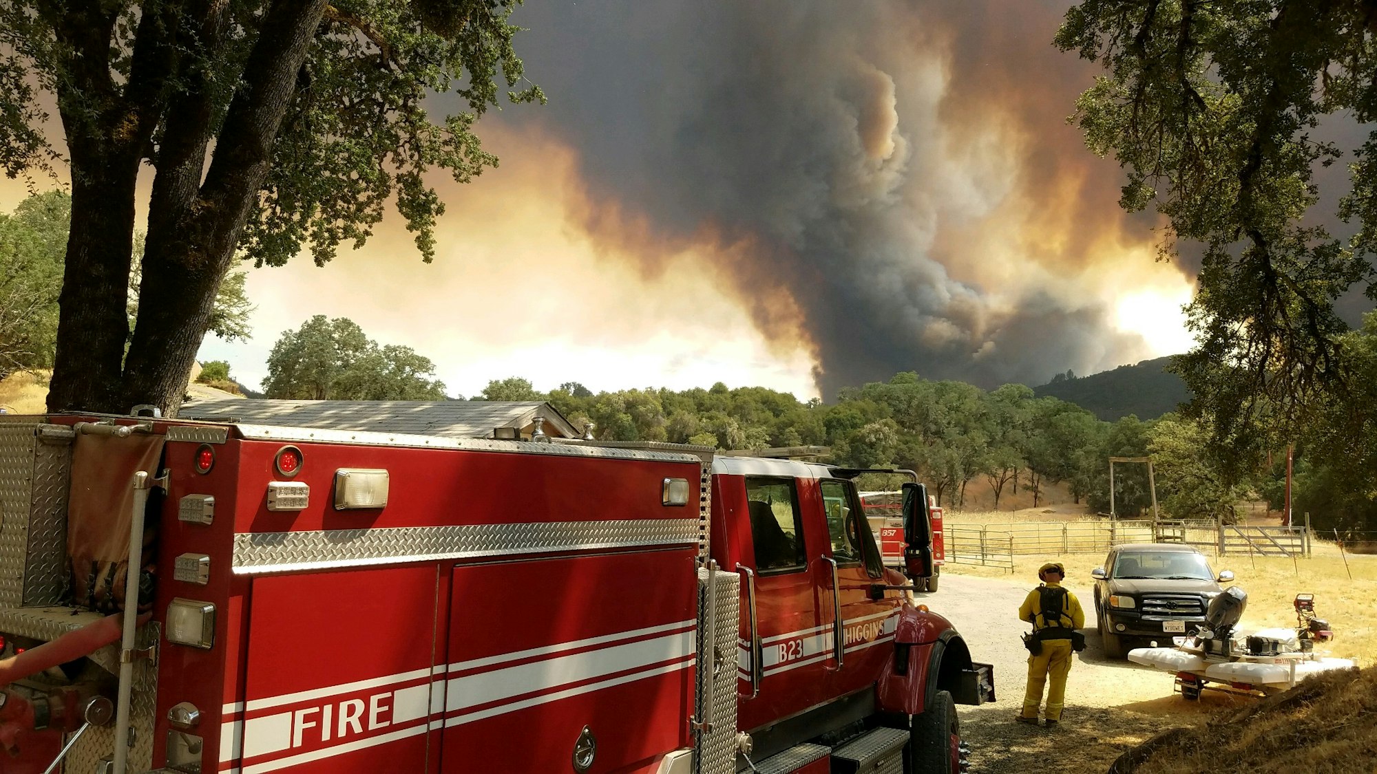 A firefighter near a fire truck with a large wildfire and smoke in the background.