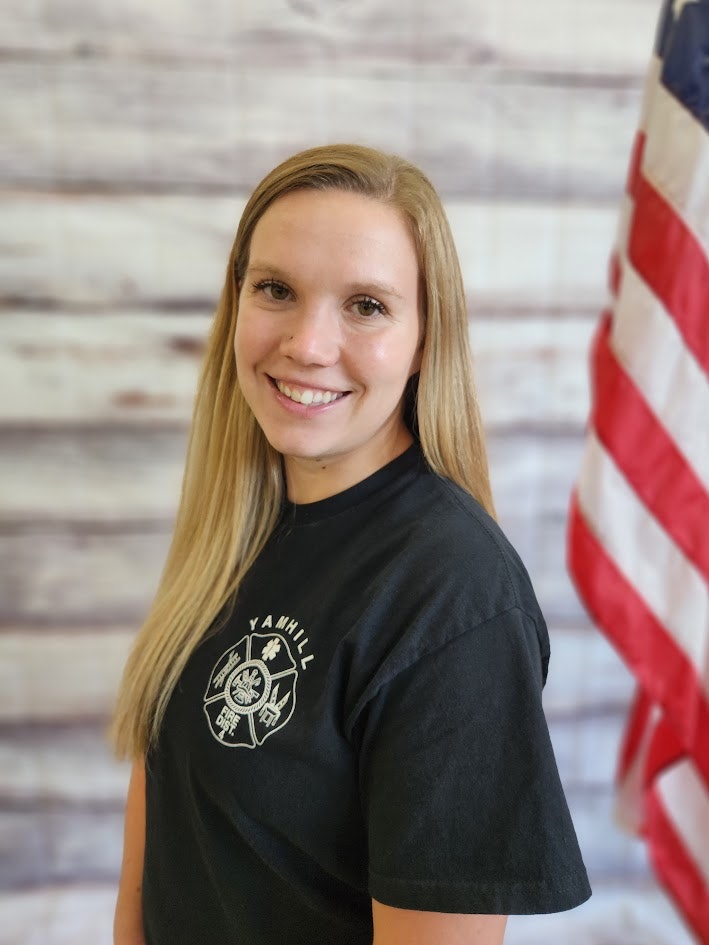 A woman smiling in a black t-shirt with a badge design, standing in front of an American flag and a wooden backdrop.