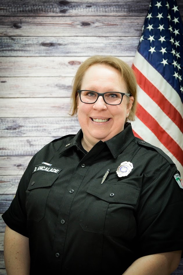 A smiling individual in a uniform with a badge, in front of a US flag and wood-patterned backdrop.
