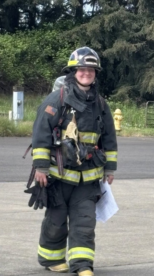 A firefighter in full gear, holding gloves and papers, standing outdoors near a hydrant.