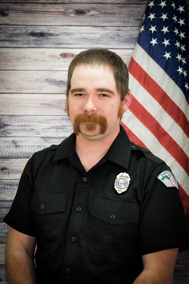 Man in uniform with badges in front of an American flag backdrop.