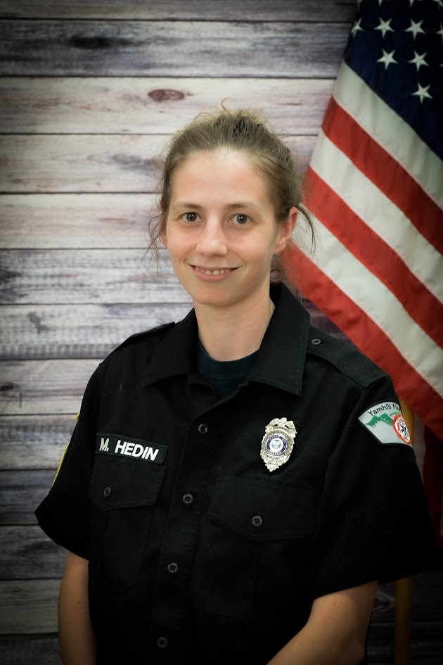 A person in a black uniform with a badge, in front of an American flag backdrop.