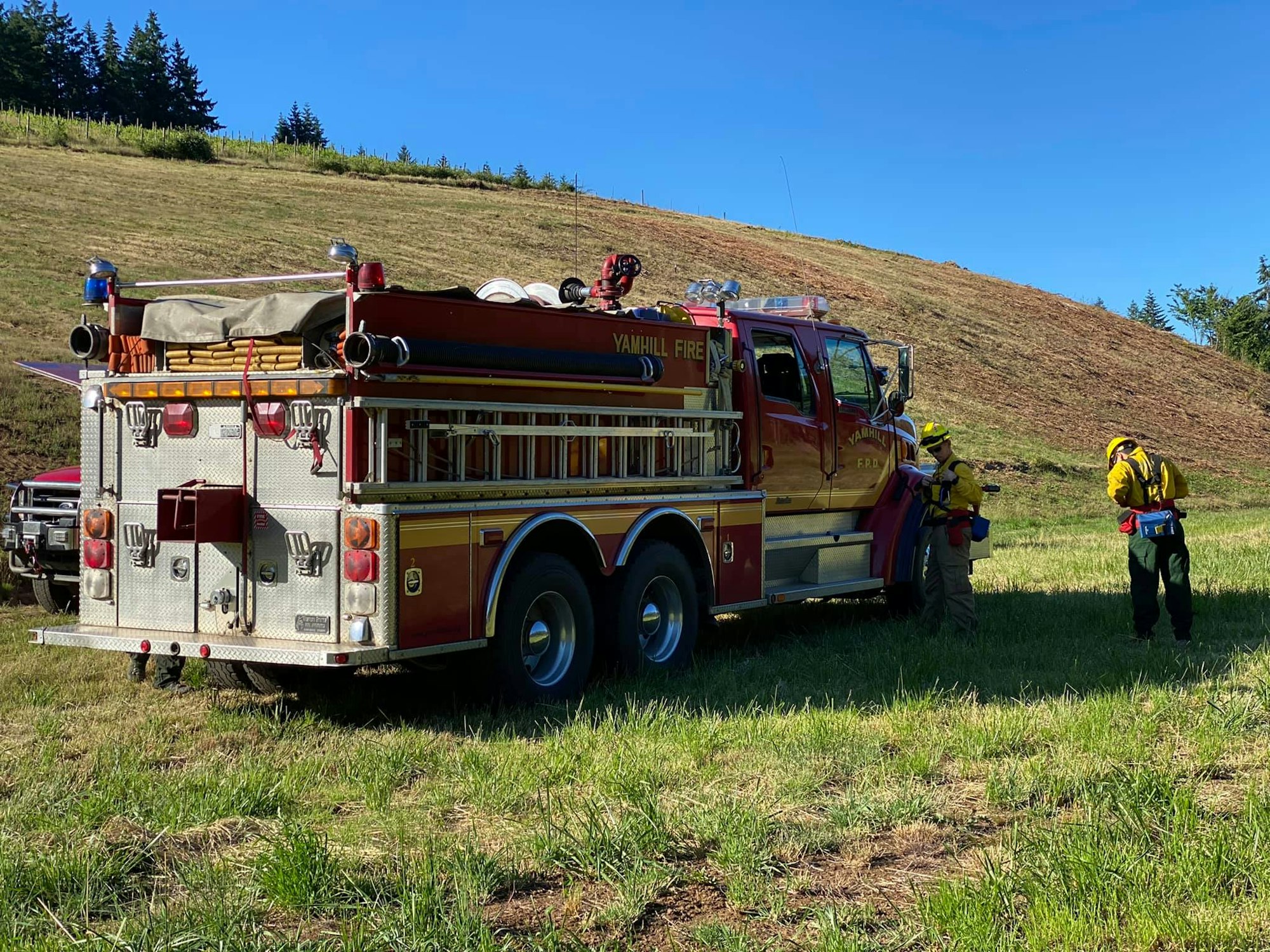 A fire truck labeled "Yamhill Fire" is parked on grassy terrain with firefighters standing nearby.