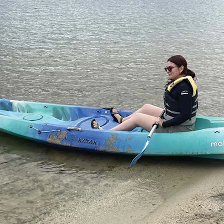 A person sitting in a kayak at the shore, wearing sunglasses and a life jacket, with calm water in the background.