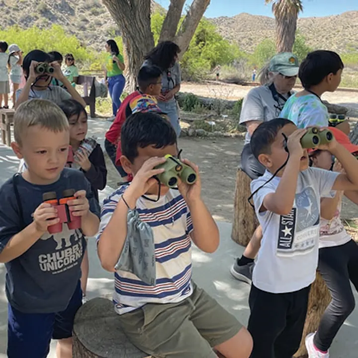 A group of children using binoculars outdoors, exploring nature while adults supervise nearby in a scenic environment.