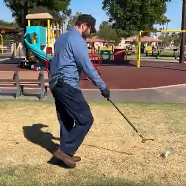 A person in a blue shirt and gloves is swinging a golf club on grass near a playground with colorful play equipment.