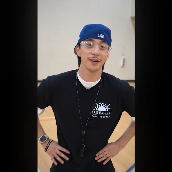 A young man wearing glasses and a cap stands in a gym, wearing a coach's shirt, looking relaxed and ready to engage.