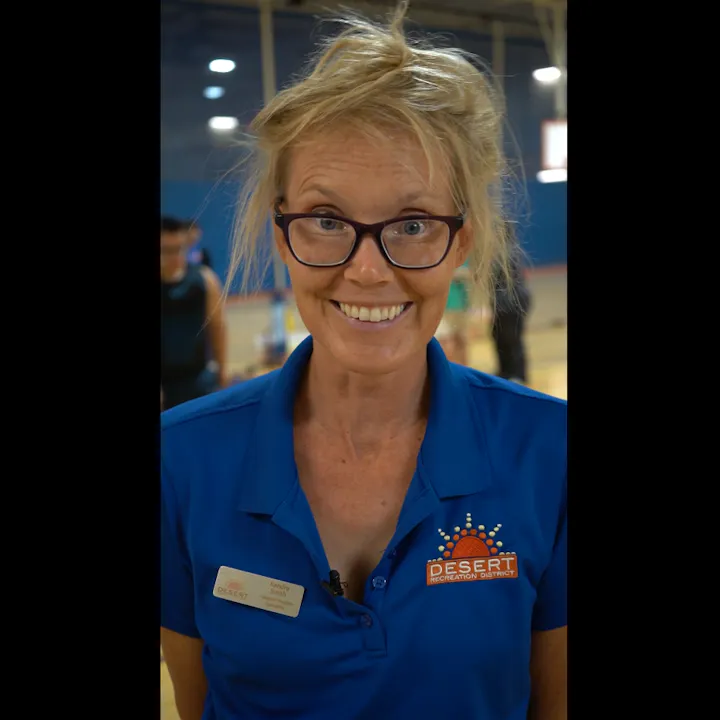 A smiling woman in a blue shirt with a "Desert Recreation District" logo, in a gym with people in the background.