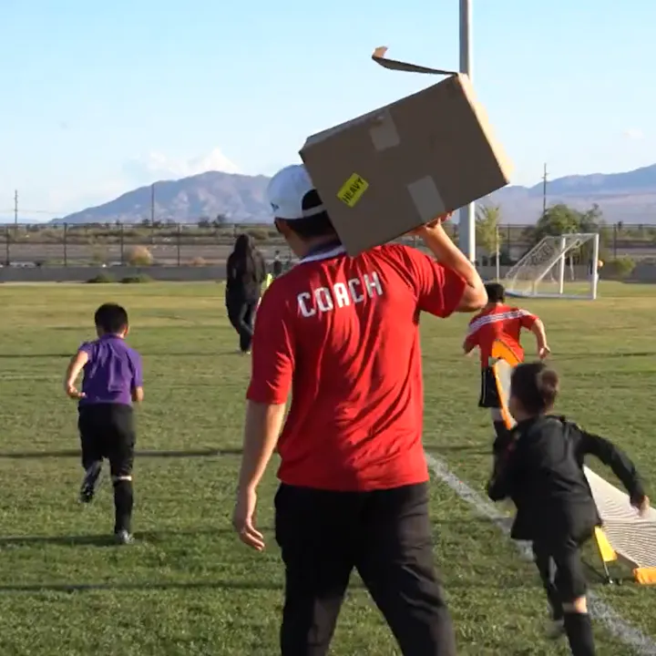 A coach in a red shirt holds a box on his shoulder while children practice soccer on a field with mountains in the background.