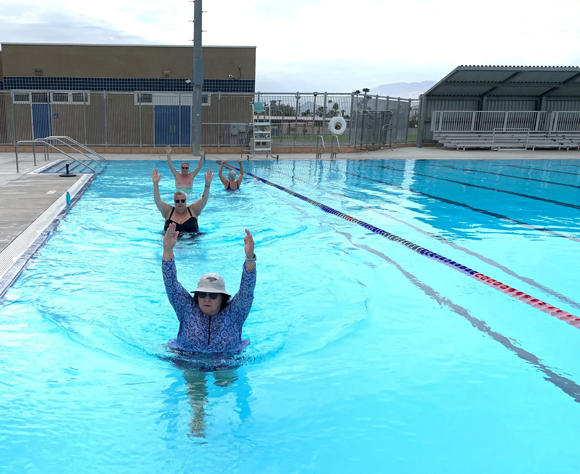 A group of people exercises in a swimming pool, with one person doing a water workout while others are swimming behind.