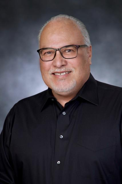 The image shows a smiling man with glasses, wearing a black shirt, against a blurred background.