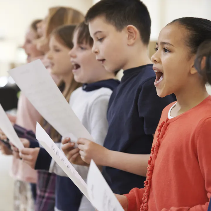A group of children singing together, holding sheet music, and expressing enthusiasm during a practice session.