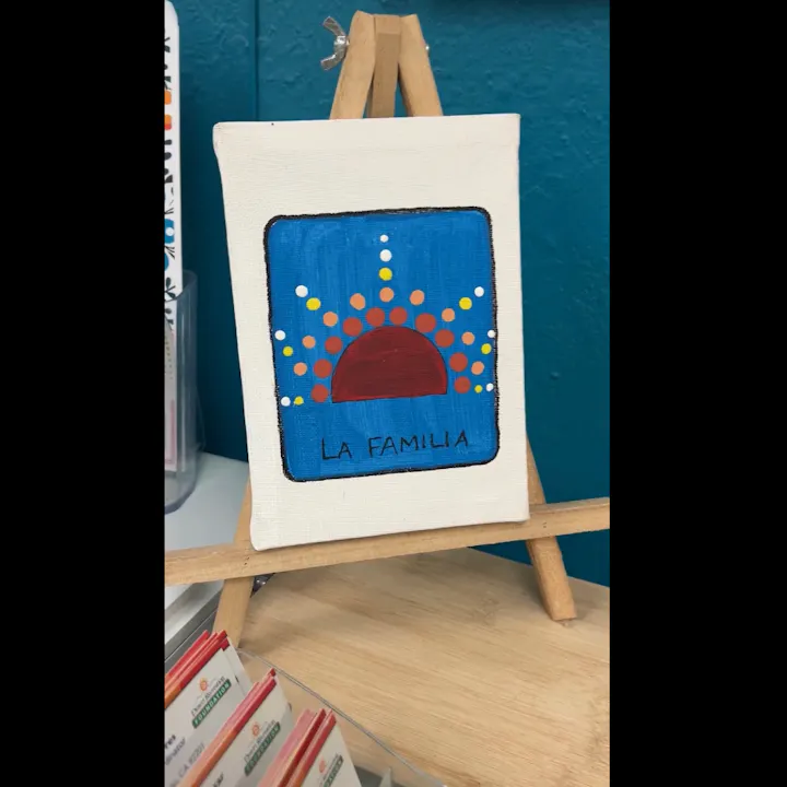 A small canvas painting featuring a sun and the words "LA FAMILIA" on a blue background, displayed on an easel.