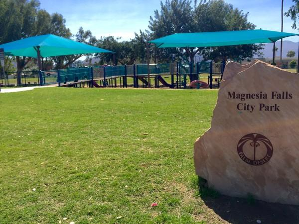 The image shows Magnesia Falls City Park, featuring green grass, playground equipment, and shaded seating areas.