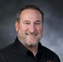 The image features a smiling man with a beard, wearing a black shirt with a logo on it, set against a neutral background.