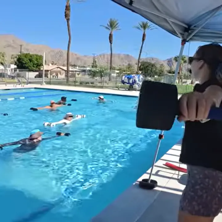 A pool setting with several people exercising in water, guided by an instructor holding a foam dumbbell under a sunny sky.
