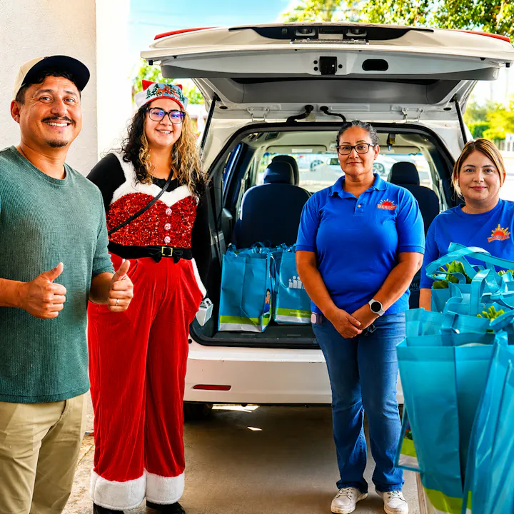 A group of four people pose by a vehicle filled with colorful bags, celebrating a festive occasion, with smiles and thumbs up.
