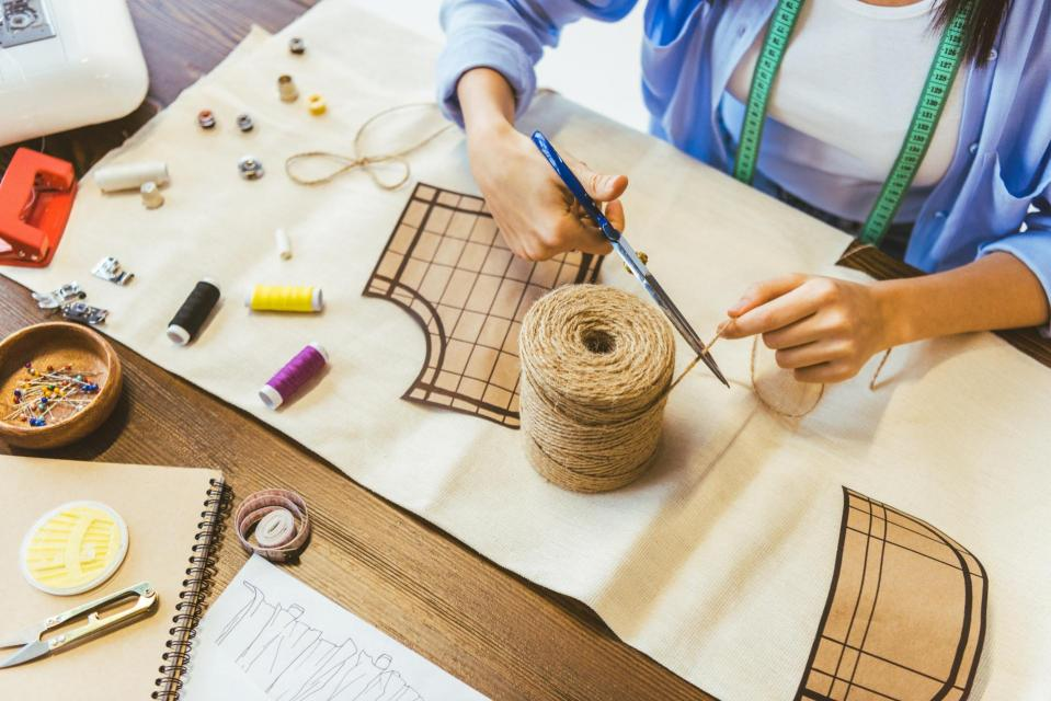 A person is cutting twine on a fabric surface, surrounded by sewing tools, threads, and a pattern layout for crafting.