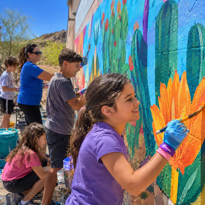 Kids and adults are painting a vibrant mural on a wall, surrounded by desert scenery and colorful flowers.