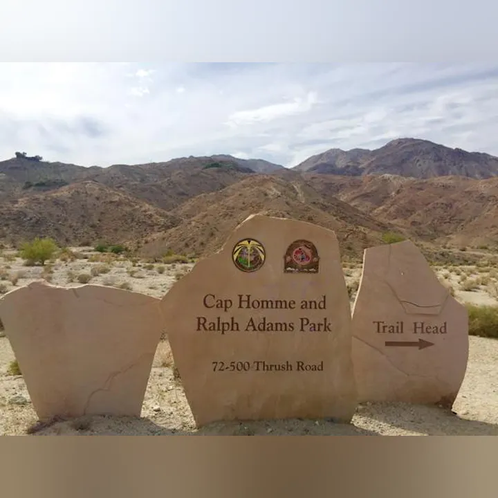 The image shows a trailhead sign for Cap Homme and Ralph Adams Park, located at 72-500 Thrush Road, surrounded by mountains.