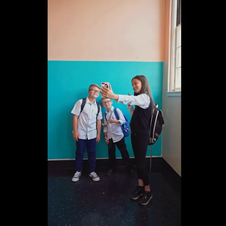 Three kids posing for a selfie in a school hallway, wearing backpacks and casual outfits, with a colorful wall in the background.