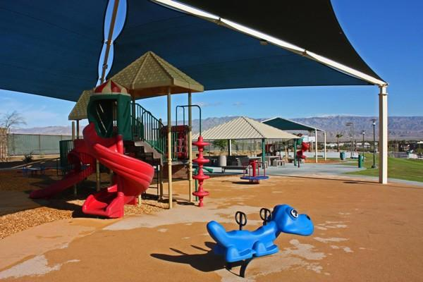 A colorful playground with a red slide, climbing structures, and a blue dinosaur rocker, all under shade canopies.
