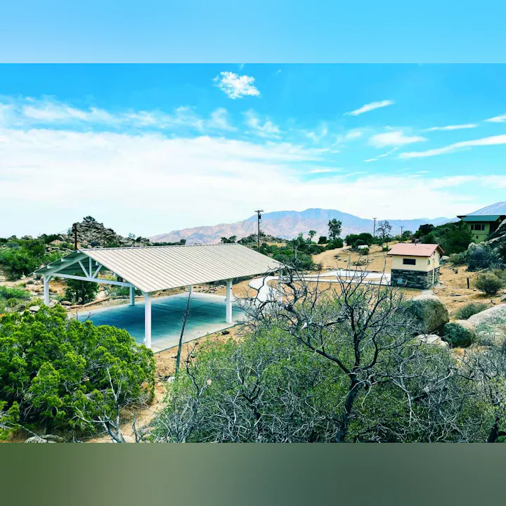 A scenic landscape featuring a metal-roofed structure, a small building, and mountain views, surrounded by desert vegetation.