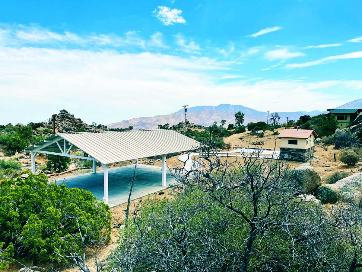 A scenic landscape featuring a metal-roofed structure, a small building, and mountain views, surrounded by desert vegetation.