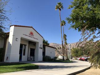 The image shows a building with palm trees, mountains in the background, and a paved pathway leading to the entrance.