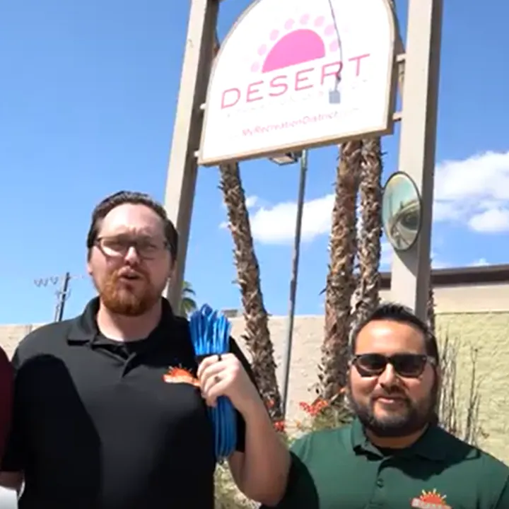 Two men standing in front of a sign for a desert recreation district, holding blue cords, with palm trees and blue sky behind them.
