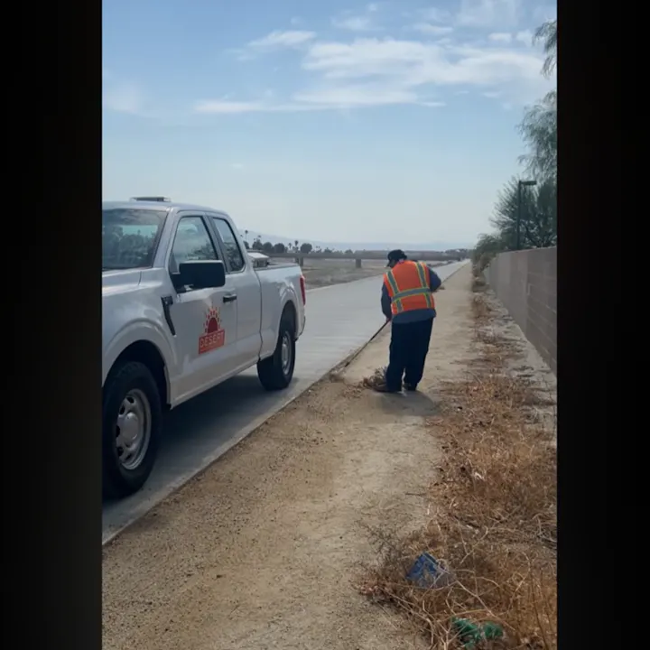 A person in an orange safety vest is picking up debris beside a road, with a white truck parked nearby.