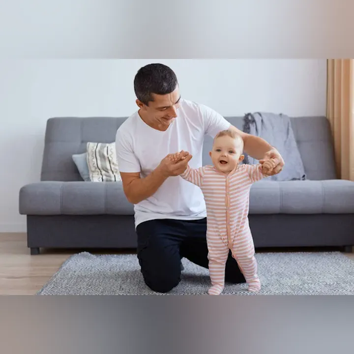 A smiling father helps his baby take steps on a rug in a cozy living room.
