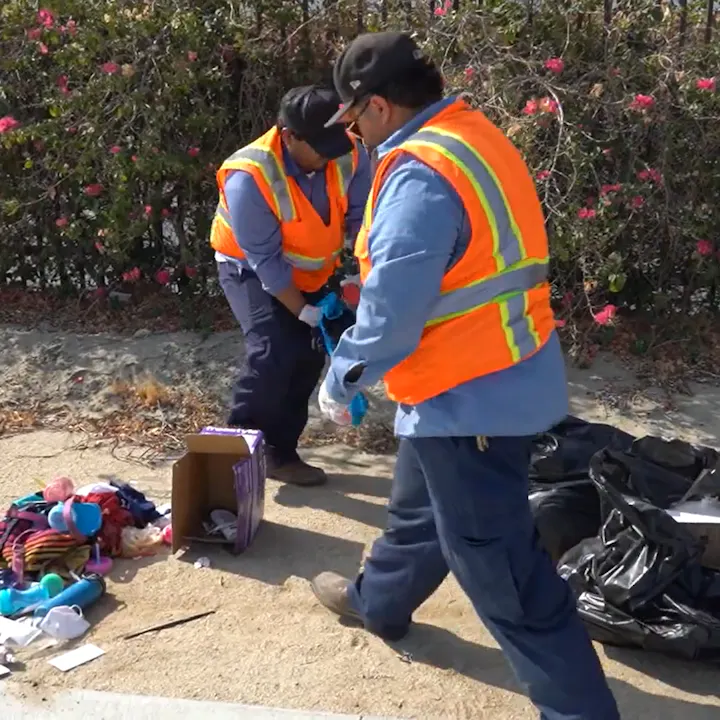 Two workers in safety vests are cleaning up trash along a roadside, sorting through items amidst flowers in the background.