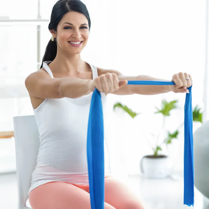 A woman exercises with a blue resistance band while sitting, smiling, in a bright room with plants.