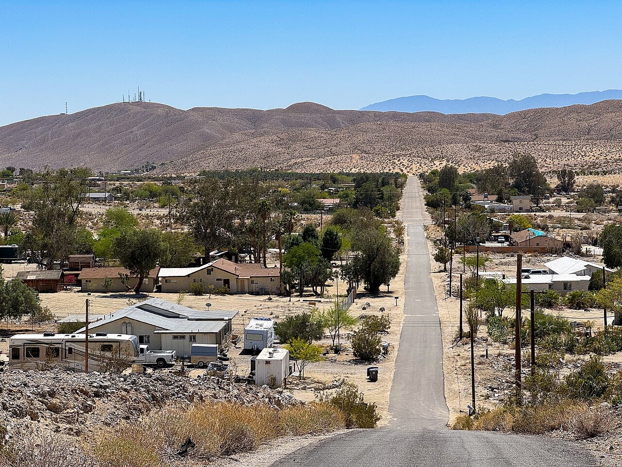 A desert landscape with a winding road, scattered homes, trees, and mountains in the background under a clear blue sky.