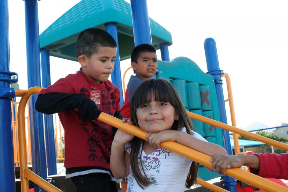 Three children play on a colorful playground, with one girl smiling as she holds onto a slide, while two boys look on.
