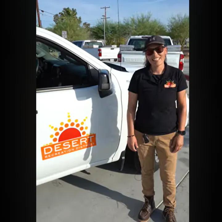 A person stands beside a white truck with a "Desert Recreation District" logo, wearing a black shirt and sunglasses.
