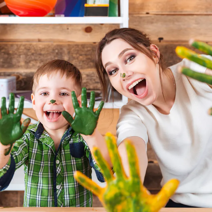 A joyful child and woman show off their paint-covered hands, smiling widely in a creative, colorful environment.