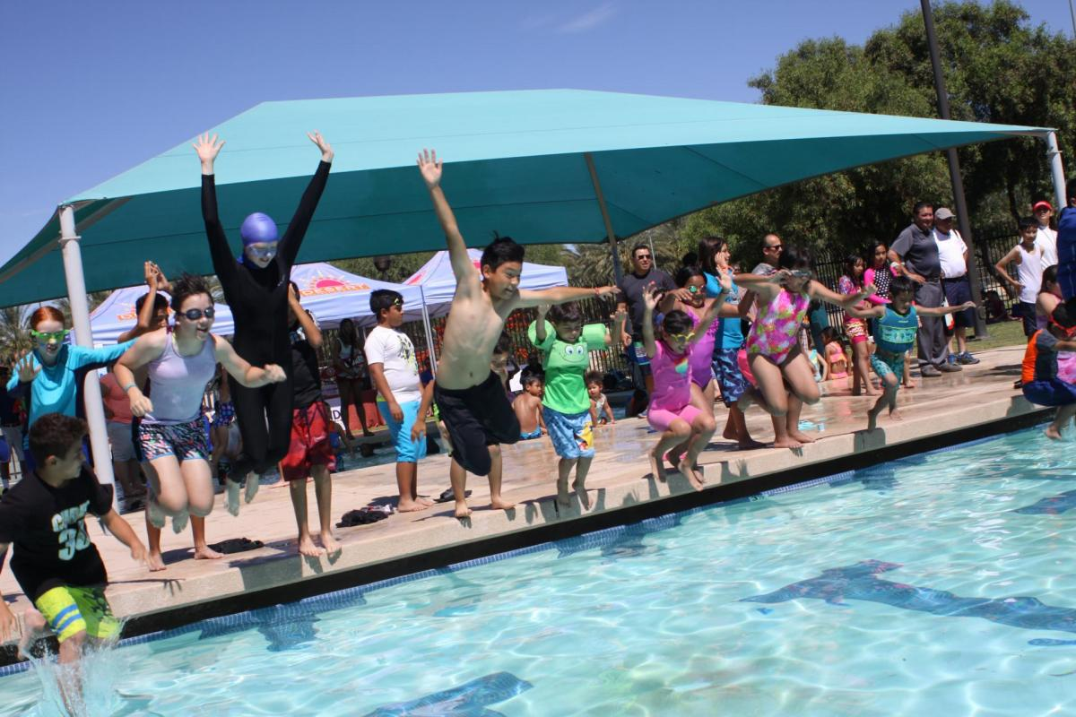 A lively scene at a pool with kids jumping in while others cheer from the side, enjoying a sunny day.