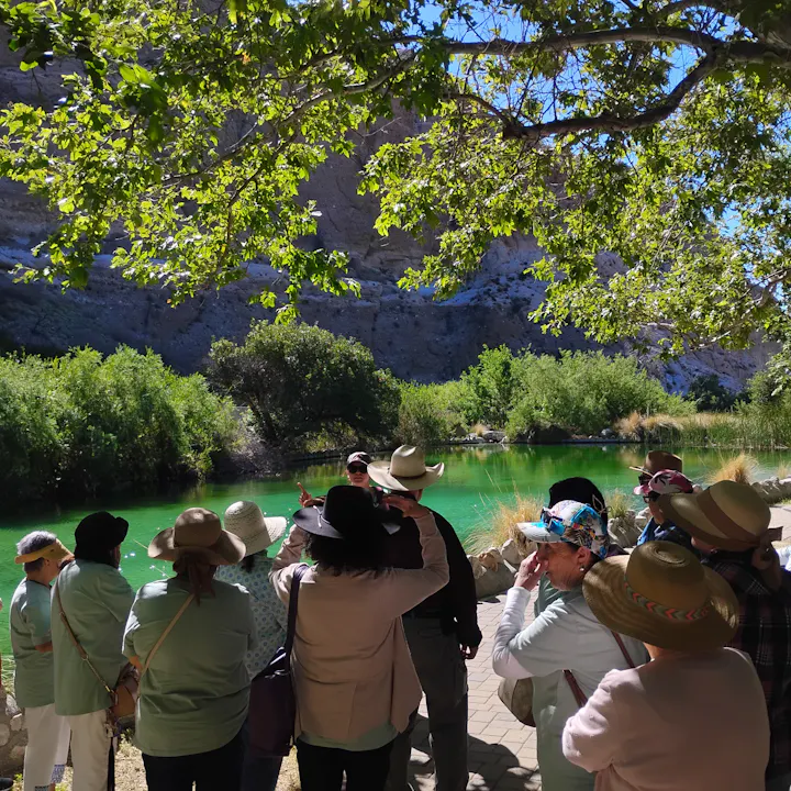 A group of people wearing hats gathered near a green pond, listening to a speaker amidst lush greenery and rocky terrain.