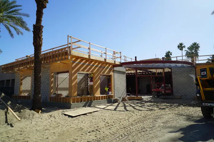 A construction site featuring a partially built house, with wooden framing, palm trees, and construction equipment in the area.