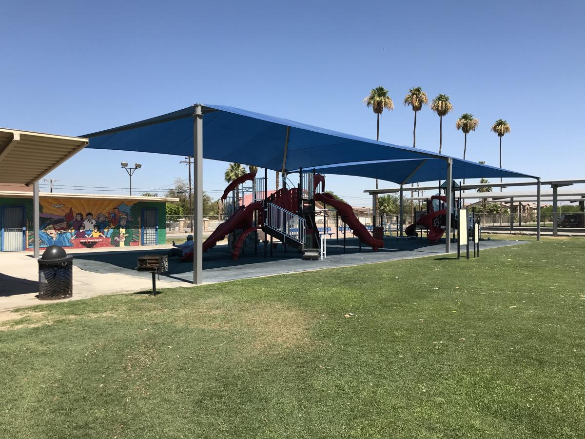 A playground with a red slide and shaded area, surrounded by green grass and palm trees, alongside a colorful mural.